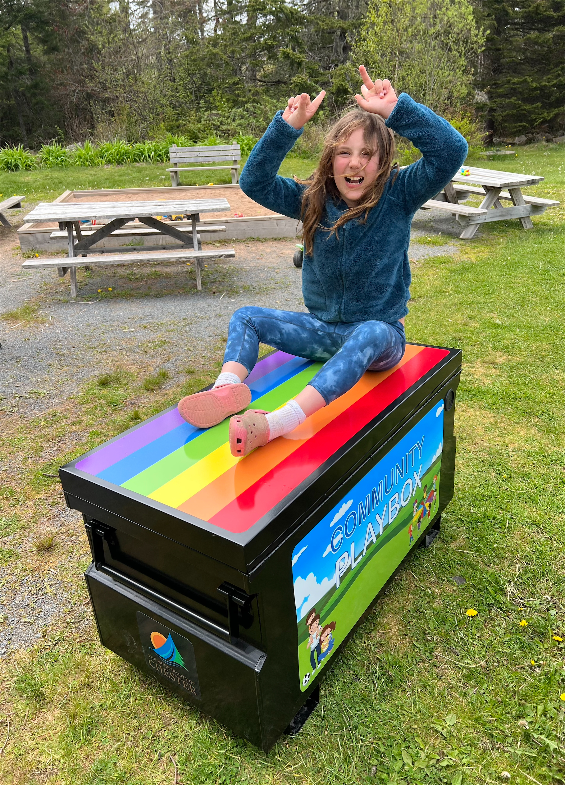A young girl with hands in the air looking excited while sitting on a brightly colored steel Playbox with toys inside.