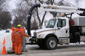 Two people in orange coveralls hold a large black hose attached to a white work truck.
