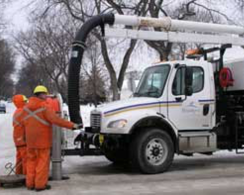 Two people in orange coveralls hold a large black hose attached to a white work truck.