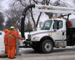 Two people in orange coveralls hold a large black hose attached to a white work truck.